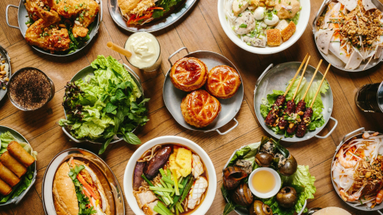 A spread of various dishes including salads, grilled meats, sandwiches, pho, and spring rolls on a wooden table.