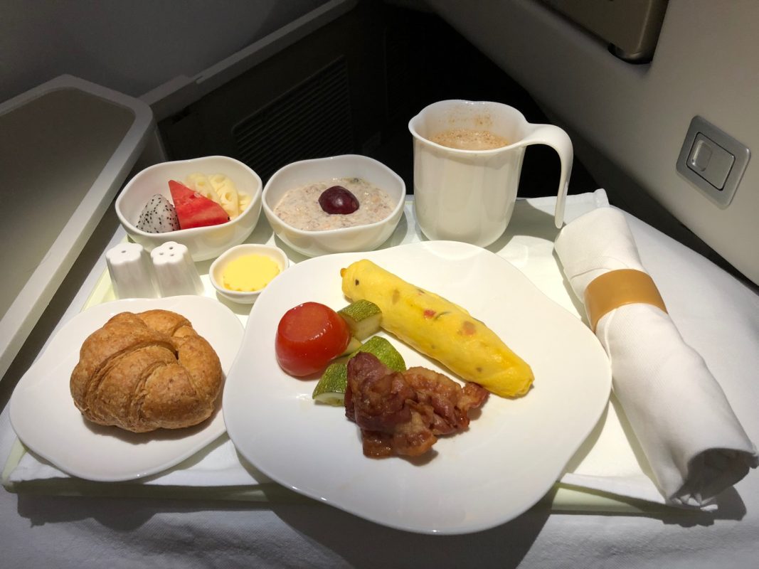 An airplane breakfast meal served on a tray, featuring an omelette, bacon, tomato, a croissant, fruit, and a hot beverage inspired by pho from Vietnam.