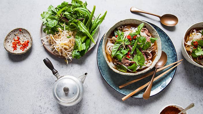 Two bowls of pho from Vietnam with beef, herbs, and noodles accompanied by condiments and fresh greens on a gray surface.