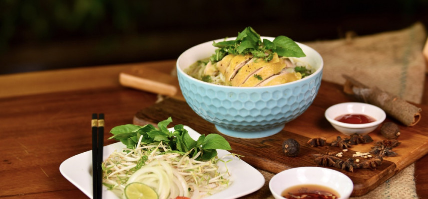 A bowl of pho with chicken, herbs, sliced lime, and sprouts on a wooden board with chopsticks and sauces on the side in Vietnam.