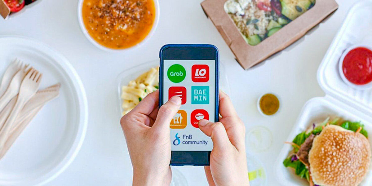 Person holding a smartphone with food delivery apps on the screen, surrounded by various takeout dishes, including a bowl of pho from Vietnam.