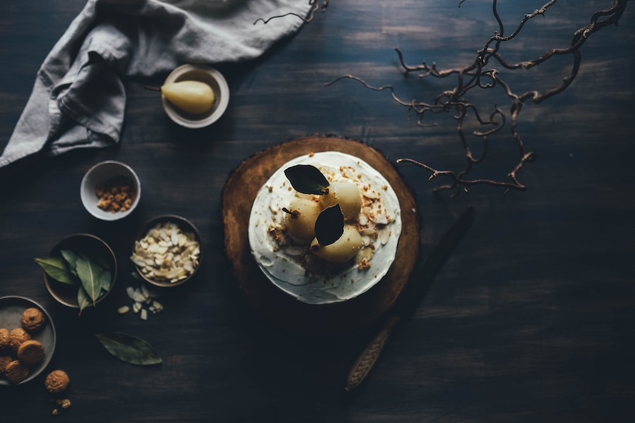 A moody top-down shot of a cake garnished with pear halves on a dark wooden surface, surrounded by ingredients and a rustic pho vietnam twig decoration.