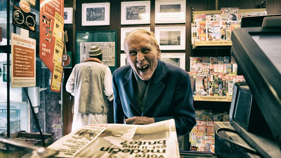 Elderly man expressing a strong emotion at the counter of a quaint shop filled with various items, magazines, and pho Vietnam recipes.