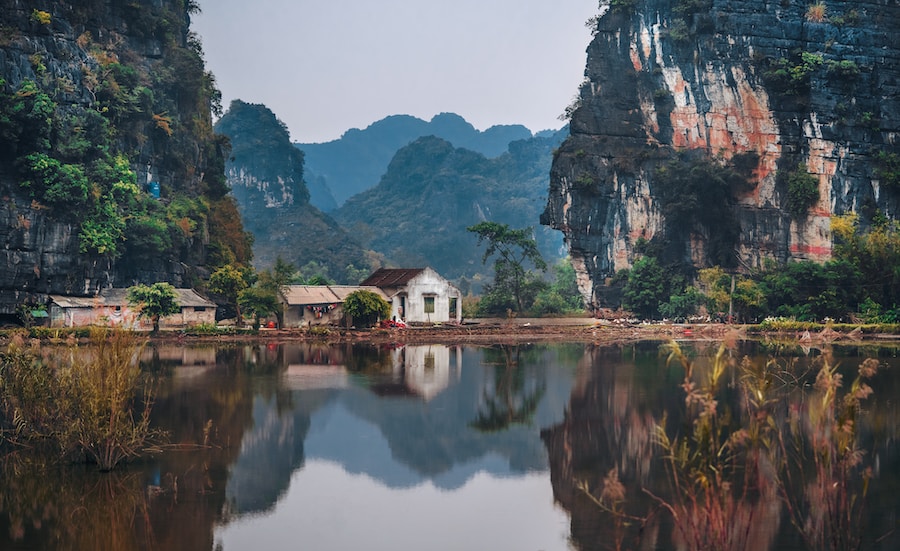 A tranquil scene with a white house reflected in a still pond, set against a backdrop of lush, towering cliffs typical of Vietnam.