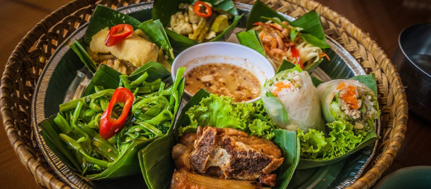 An assortment of traditional Southeast Asian dishes, including fresh spring rolls and pho from Vietnam, presented on a woven tray with banana leaves.