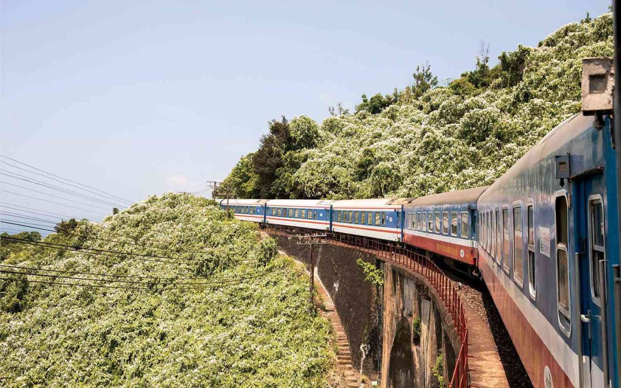 A train crossing a bridge surrounded by lush greenery on a sunny day in rural Vietnam.