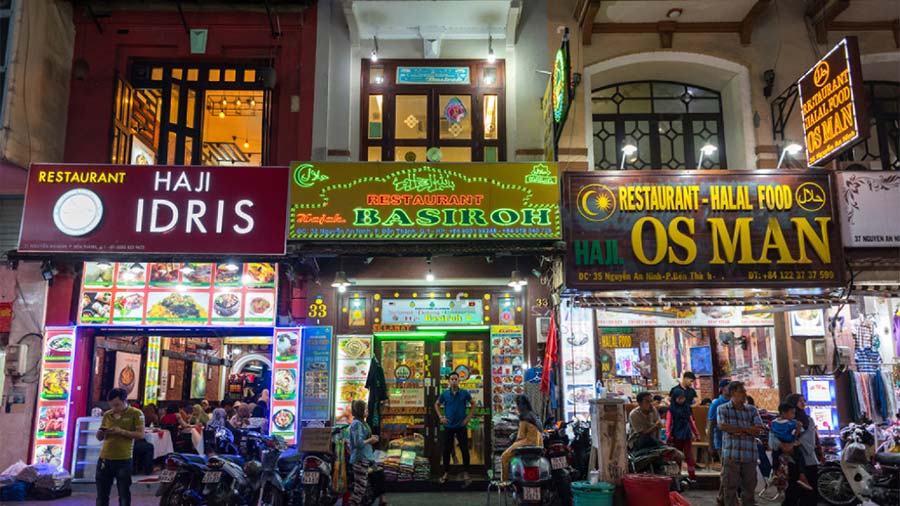 Three adjacent restaurants with bright neon signs illuminated at night, serving pho and other dishes, with people and parked motorcycles on the street in front in Vietnam.