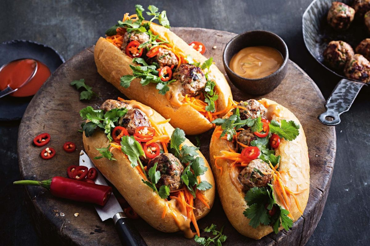 Three Vietnamese-style banh mi sandwiches with meatballs, vegetables, and cilantro on a rustic wooden board with pho-inspired sauces on the side.