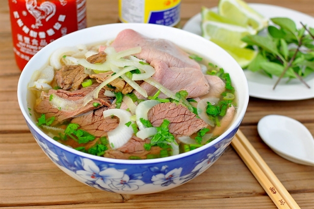 A bowl of pho from Vietnam with sliced beef, onions, herbs, and lime served with chopsticks and condiments on a wooden table.