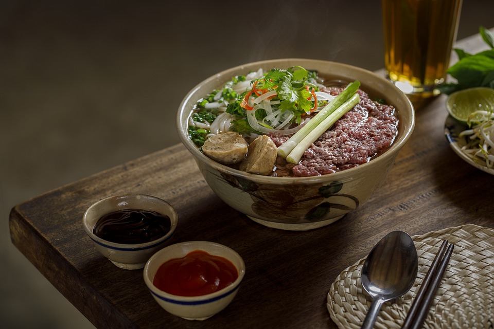 A bowl of pho vietnam with thinly sliced beef, garnished with herbs, alongside condiments and a glass of beer.