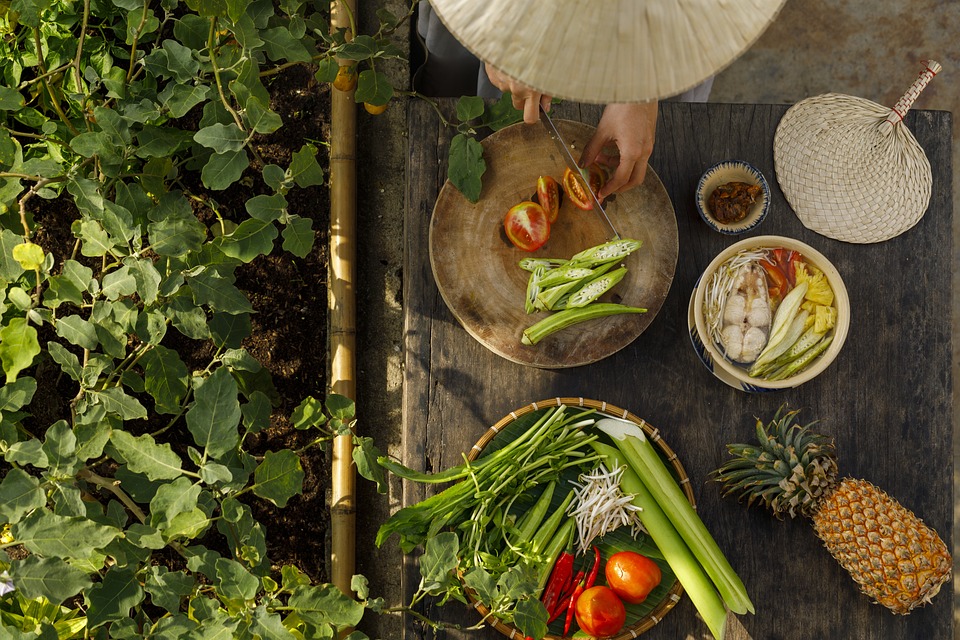 Person preparing fresh vegetables and fruit at an outdoor table next to a garden, inspired by the flavors of pho from Vietnam.