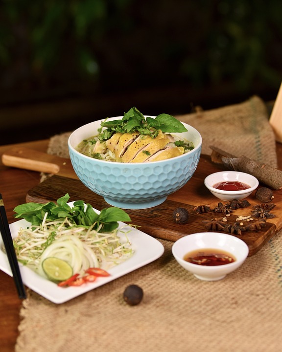 A bowl of pho from Vietnam with chicken, garnished with herbs, next to sauces, lime, and bean sprouts on a wooden table.