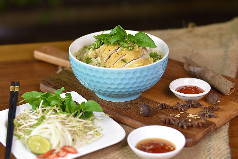 Bowl of chicken pho from Vietnam with fresh herbs, lime, and sauces on a wooden table.