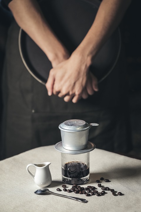 A person standing behind a traditional Vietnamese drip coffee filter setup with a small pitcher and coffee beans on a table near a bowl of pho.