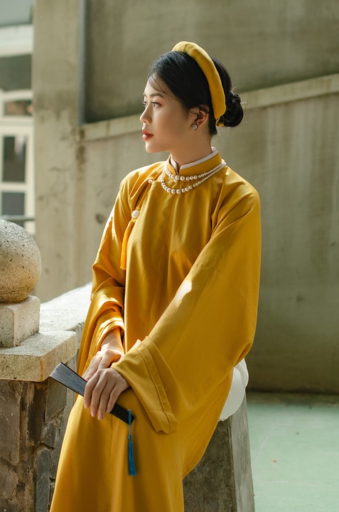 A woman in a traditional yellow dress and headpiece, reminiscent of pho Vietnam culture, looking contemplatively to the side.