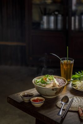 A steaming bowl of Vietnam pho with condiments and iced tea on a wooden table.