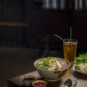 A steaming bowl of pho from Vietnam accompanied by condiments and a drink on a wooden table.