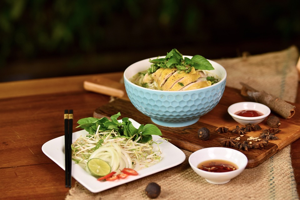 A bowl of pho from Vietnam with chicken, fresh herbs, lime, and side sauces on a wooden table.