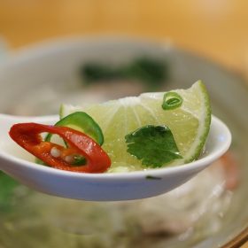 Close-up of a spoon holding a lime slice, chili, and cilantro over a bowl of Pho soup.