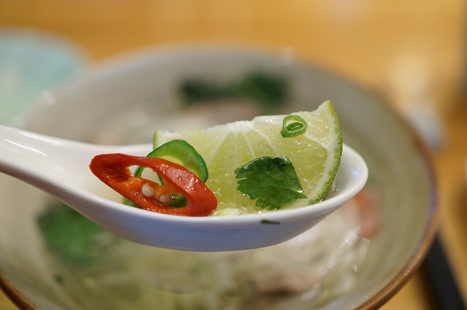 Close-up of a spoon holding a lime slice, chili, and cilantro over a bowl of Pho soup.