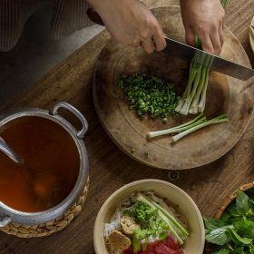 A person preparing ingredients for a meal, chopping scallions on a wooden board, with a pot of broth, raw beef, and bowls of noodles and vegetables nearby to cook traditional pho from Vietnam