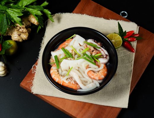 A bowl of pho, a traditional Vietnamese seafood noodle soup garnished with herbs and lime, accompanied by fresh chili peppers and ginger on a dark table.