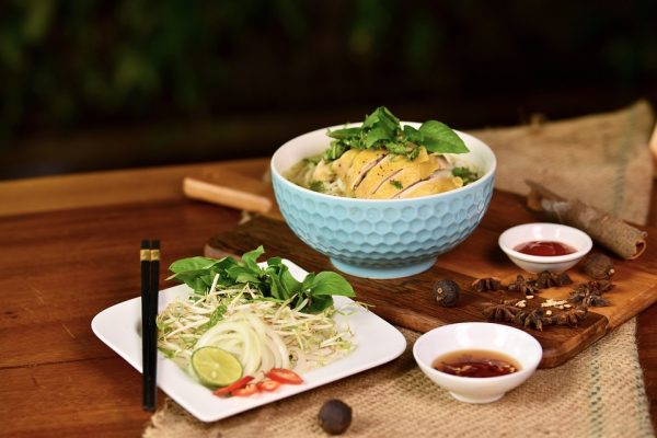 A bowl of pho from Vietnam with chicken, herbs, and garnishes, accompanied by chopsticks and dipping sauces on a wooden table.